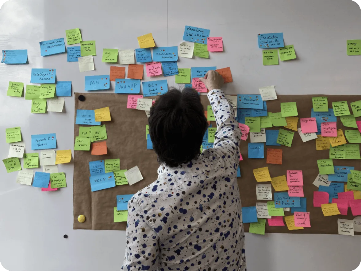 A woman writing on a bulletin board covered in sticky notes.